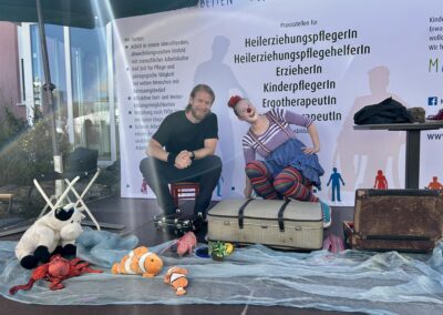 Ein Mann und eine als Clown verkleidete Frau sitzen auf einer Bühne und lachen in die Kamera.